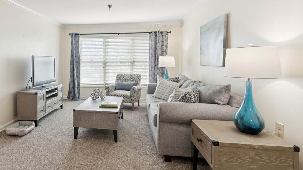 A bright living room with a large window, light gray sofa, armchair, two wooden tables, TV on a stand, blue glass lamp, neutral décor, and soft carpet flooring.