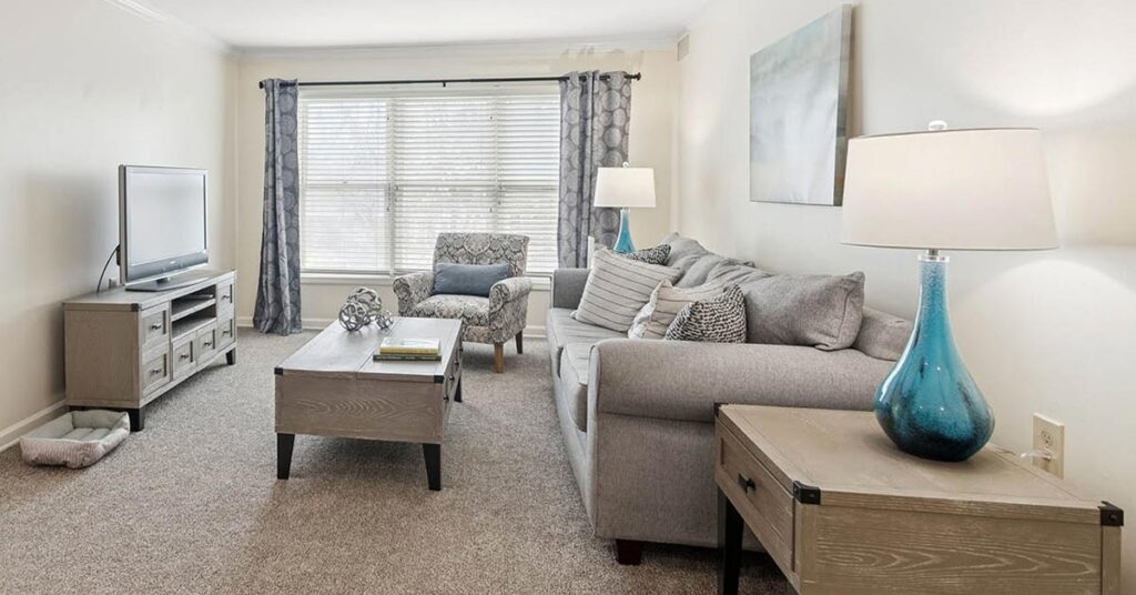 A bright, modern living room with a gray sofa, patterned armchair, wooden coffee tables, TV on a stand, large window with curtains, and a blue glass table lamp. Neutral tones and minimal décor create a cozy atmosphere.