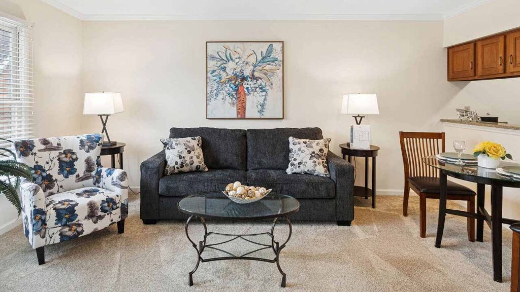 A cozy living room with a dark gray sofa, two patterned pillows, a floral armchair, glass coffee table, two lamps, a round dining table with two chairs, and a large floral painting on a cream wall.