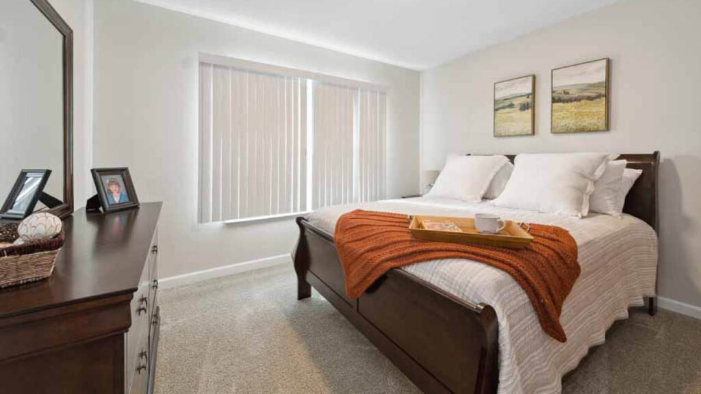 A cozy bedroom with a large bed featuring white bedding, an orange knit blanket, and a tray with a cup. There are two pictures above the bed, a dresser with framed photos, and vertical blinds covering a window.
