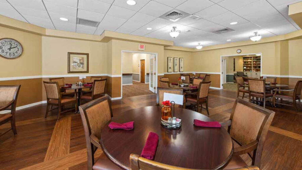 A spacious dining room with wooden tables and chairs, set with red napkins. Warm tan and beige walls, framed artwork, a clock, and ceiling lights create a welcoming atmosphere. An open doorway leads to a bookshelf area.