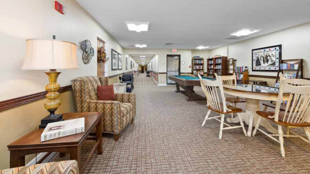 A spacious, well-lit common room with armchairs, a lamp, a side table, bookshelves, a pool table, and a round table with chairs. The walls are decorated with art and the carpeted floor extends down a hallway.