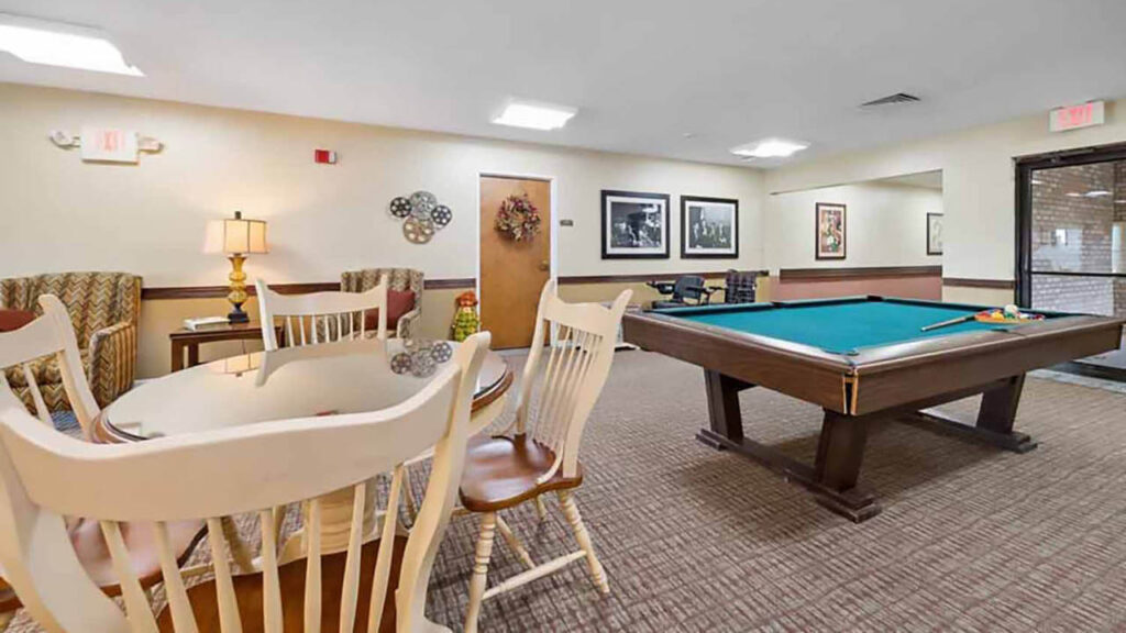 A recreation room with a pool table, cue sticks, a round dining table with chairs, two armchairs, a side table with a lamp, wall decor, framed photos, and neutral-toned carpet flooring.