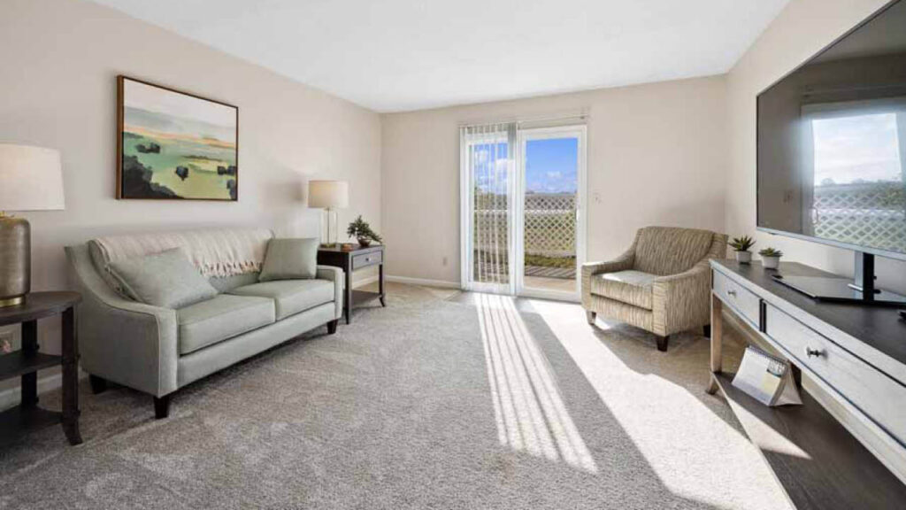 Bright living room with beige carpet, a light gray sofa, an armchair, a TV on a stand, a side table with a lamp, wall art, and glass doors leading to a balcony with a view of the outdoors.
