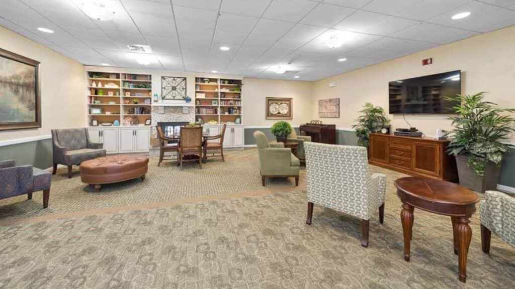 A spacious, well-lit lounge with armchairs, tables, a sofa, a wall-mounted TV, bookshelves, and decorative plants. The room has patterned carpet and neutral-colored walls with framed artwork.