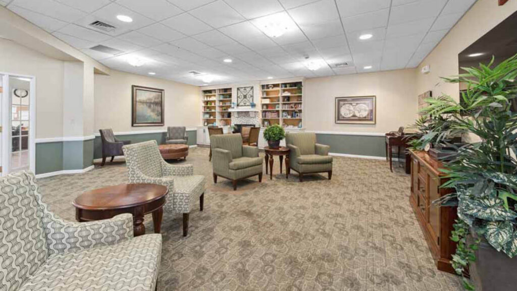 A spacious, well-lit lounge with patterned armchairs, round wooden tables, a built-in bookshelf, artwork on beige walls, and decorative plants, creating a cozy and inviting atmosphere.