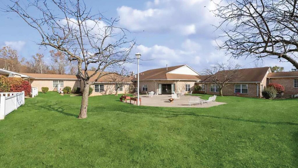 A large, single-story brick building with a brown roof is surrounded by a green lawn, leafless trees, and patio furniture under a partly cloudy sky.