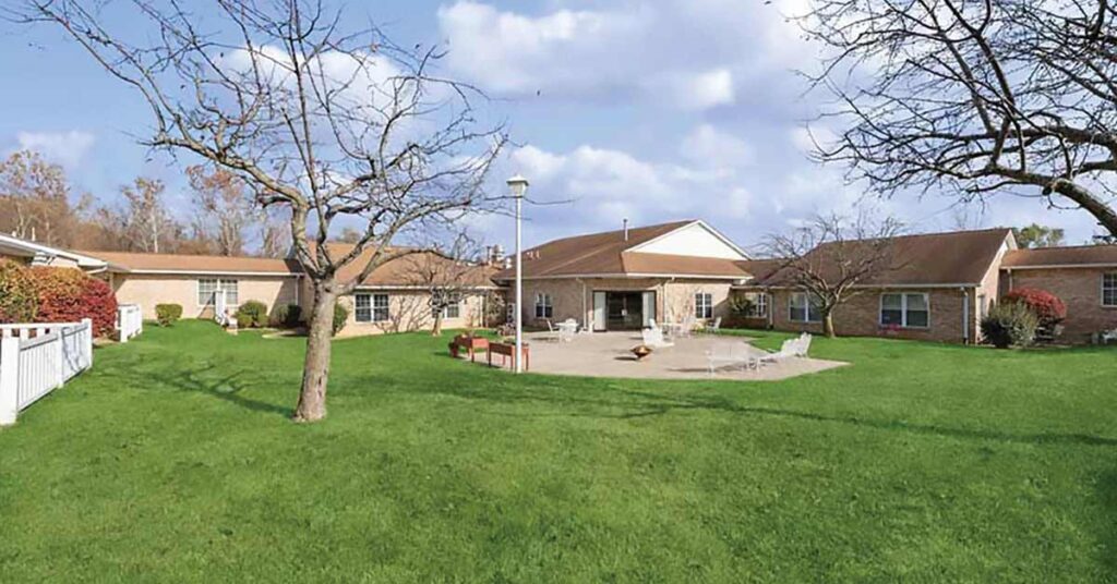 A wide view of a single-story brick building with a brown roof, surrounded by green grass, leafless trees, and outdoor seating on a patio under a partly cloudy sky.
