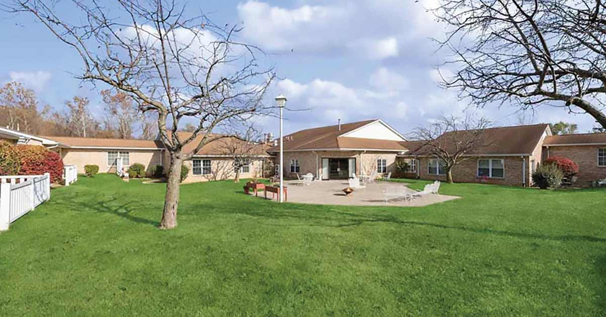 A wide view of a single-story brick building with a brown roof, surrounded by green grass, leafless trees, and outdoor seating on a patio under a partly cloudy sky.