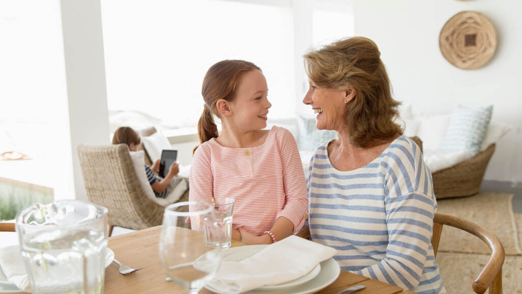A smiling older woman and a young girl sit together at a dining table, facing each other. In the background, another child sits on a chair using a tablet. The room is bright and cozy with light decor.