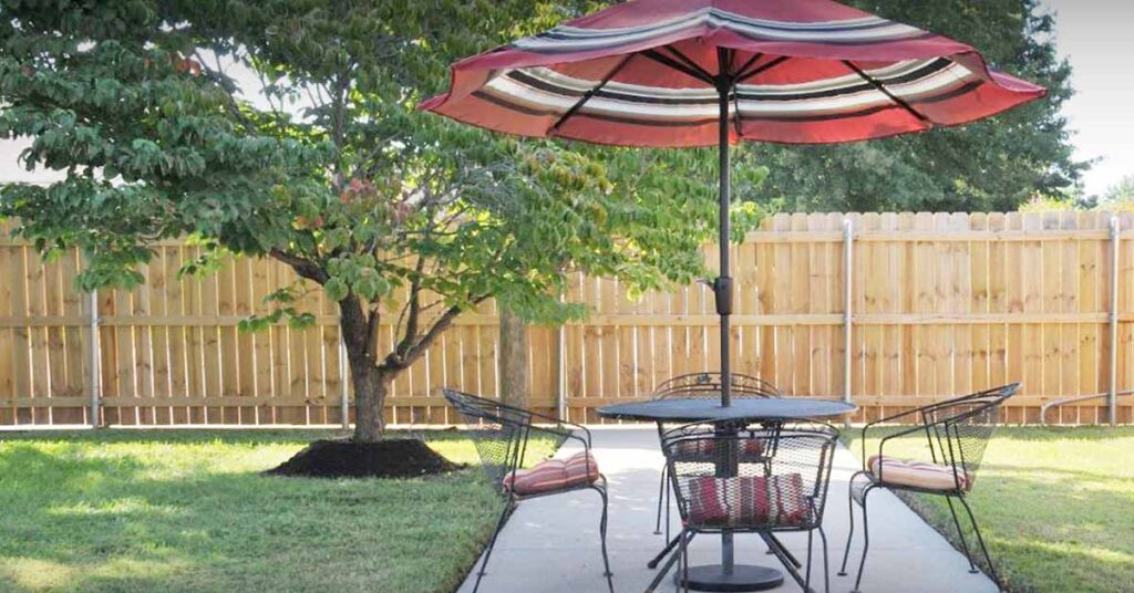 A patio with a round table and four chairs sits under a large red umbrella on a concrete path. There is green grass, a tree, and a wooden fence in the background.