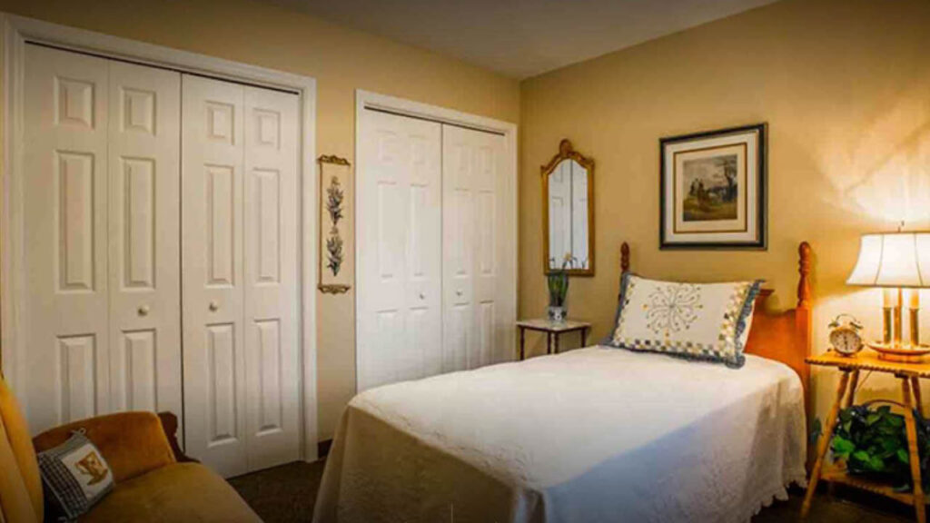 A cozy bedroom with a neatly made single bed, a white quilt, decorative pillows, two white double-door closets, a mirror, framed artwork, a lamp, and a small clock on a wooden nightstand.