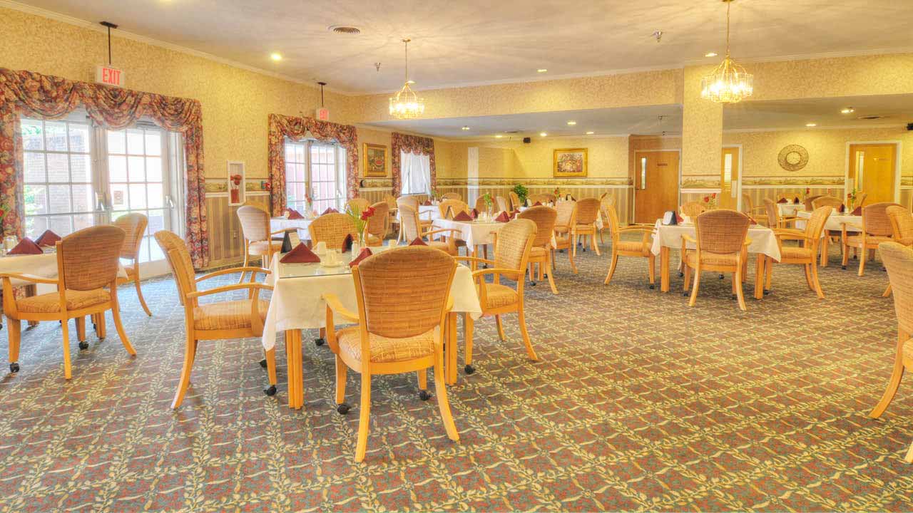 A brightly lit dining room with several tables set with white tablecloths and napkins, surrounded by wooden chairs. The room has large windows, patterned carpet, and chandeliers hanging from the ceiling.