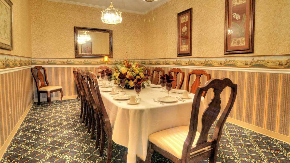 A formal dining room with a long table set for a meal, surrounded by wooden chairs. The room features patterned wallpaper, floral carpet, a mirror, framed art, and a chandelier overhead.