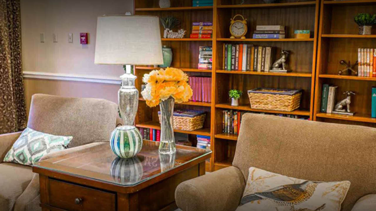 Two beige armchairs with patterned pillows sit beside a wooden side table holding a lamp and a vase of yellow flowers. In the background, a bookshelf is filled with books, baskets, and decorative items.