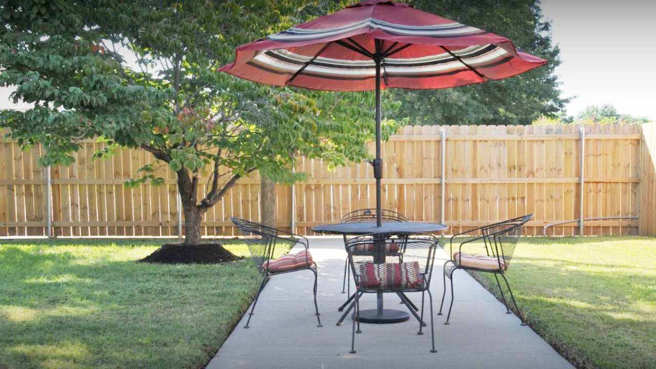 A patio set with four metal chairs and a round table sits on a concrete path in a backyard. A large red umbrella provides shade, and a wooden fence and leafy tree are in the background.