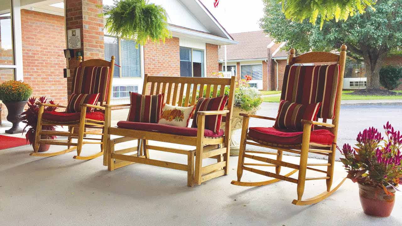 Two wooden rocking chairs and a matching bench with red and striped cushions sit on a covered porch. Potted plants and hanging ferns decorate the area, with brick buildings and trees in the background.