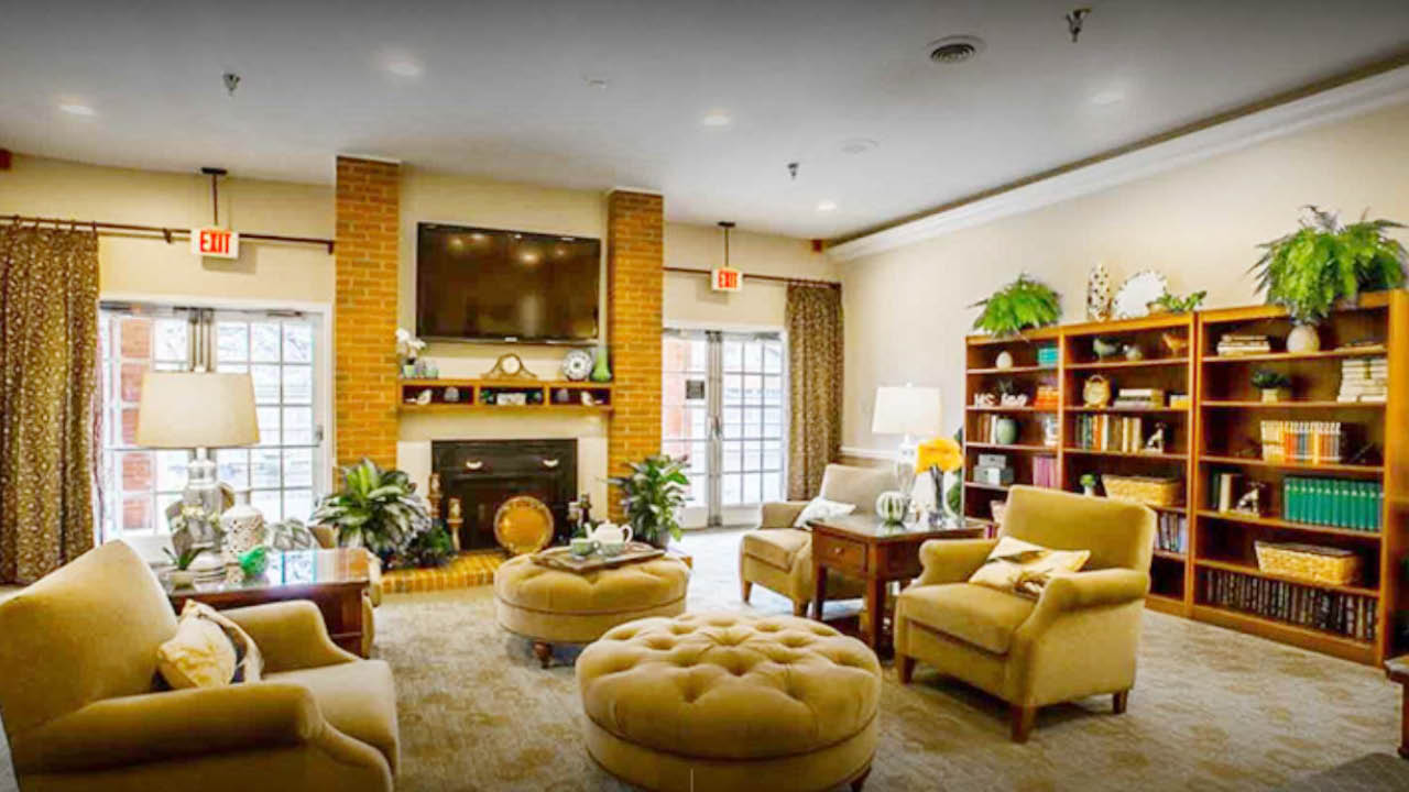 A cozy living room with beige armchairs, a large round ottoman, a fireplace beneath a wall-mounted TV, bookshelves filled with books, potted plants, and large windows with patterned curtains.
