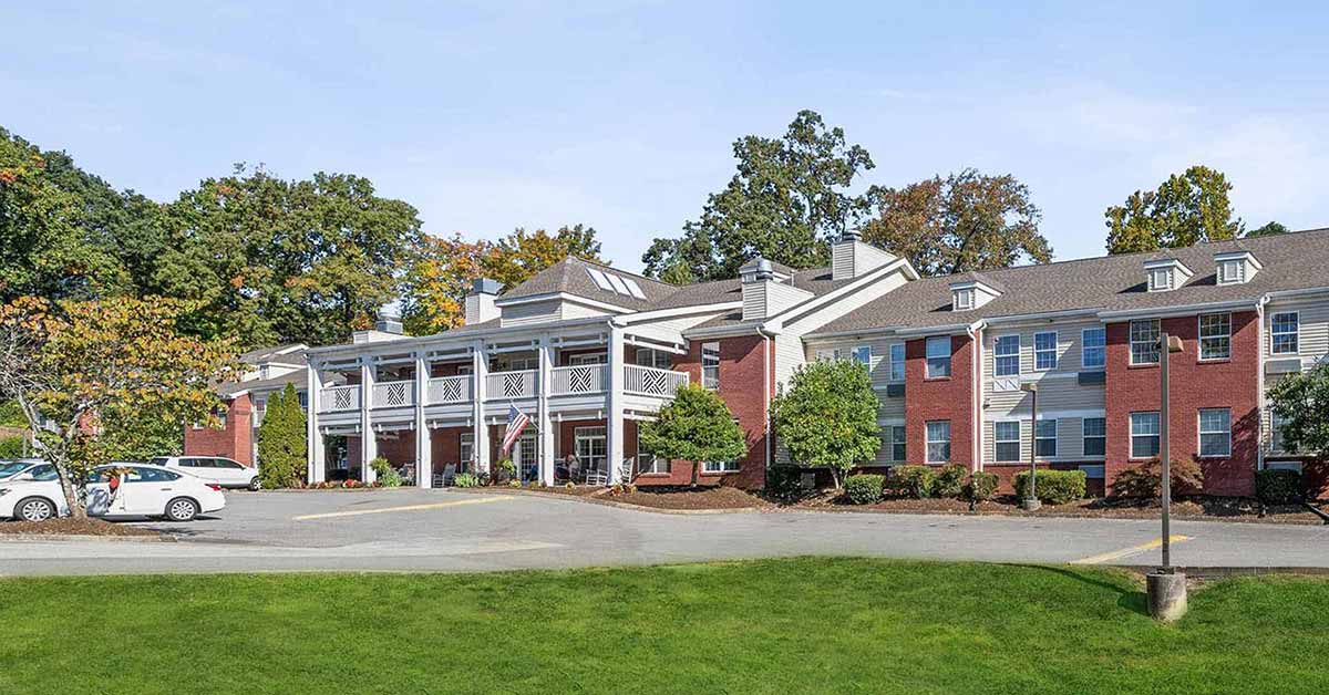 A large, two-story brick building with white columns and balconies, surrounded by trees and greenery. Several cars are parked in front, and the sky is clear and blue.