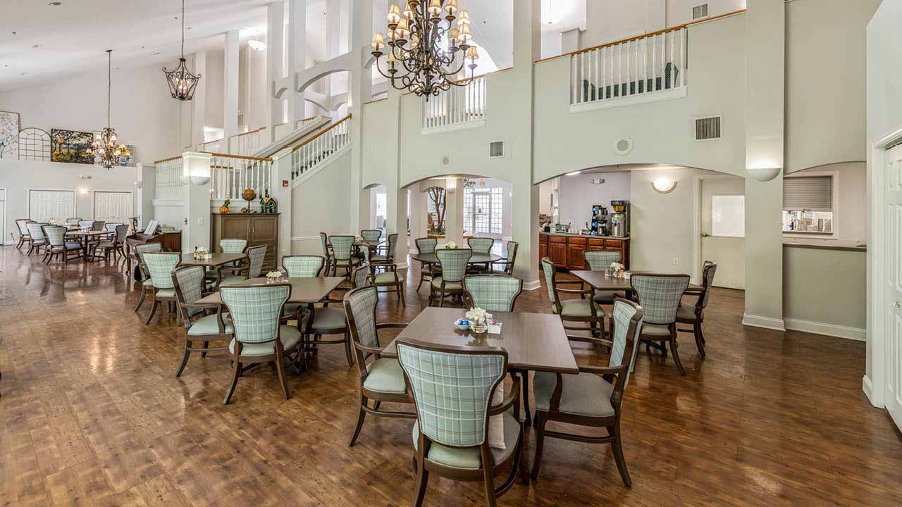 A spacious, elegant dining area with wooden floors, light-colored cushioned chairs, square tables, high ceilings, chandeliers, and large windows letting in natural light. The room features a split-level balcony and a coffee station.