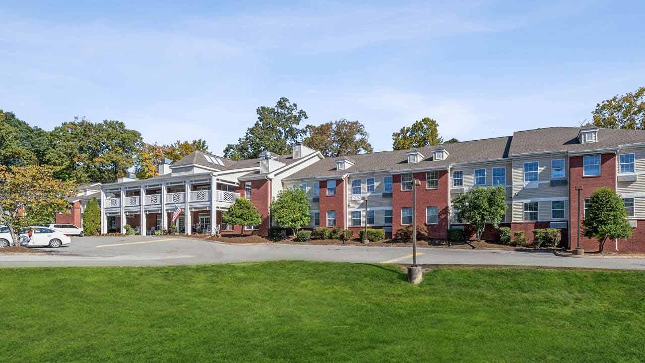 A large red-brick building with white columns and balconies, surrounded by trees and greenery, with parked cars in front and a clear blue sky overhead.