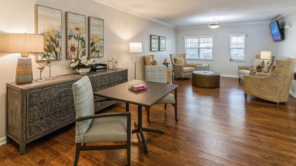 A cozy, well-lit living room with wooden floors, armchairs, a patterned ottoman, a desk with chairs, decorative lamps, floral artwork, and large windows letting in natural light.