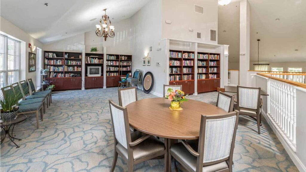 A bright, spacious room with bookshelves along the back wall, a row of chairs beside large windows, and a round table with six chairs in the foreground. A flower arrangement sits on the table.