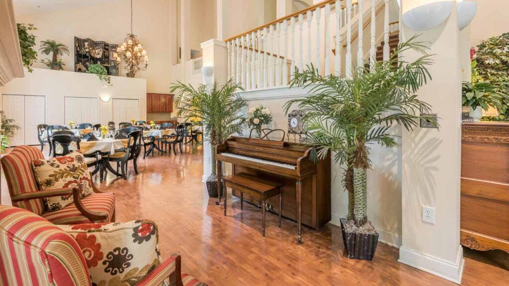 A spacious, well-lit room with a piano, two striped armchairs, potted plants, and a dining area set with tables and chairs. A staircase leads to an upper floor, and the space features wooden floors and decorative lighting.