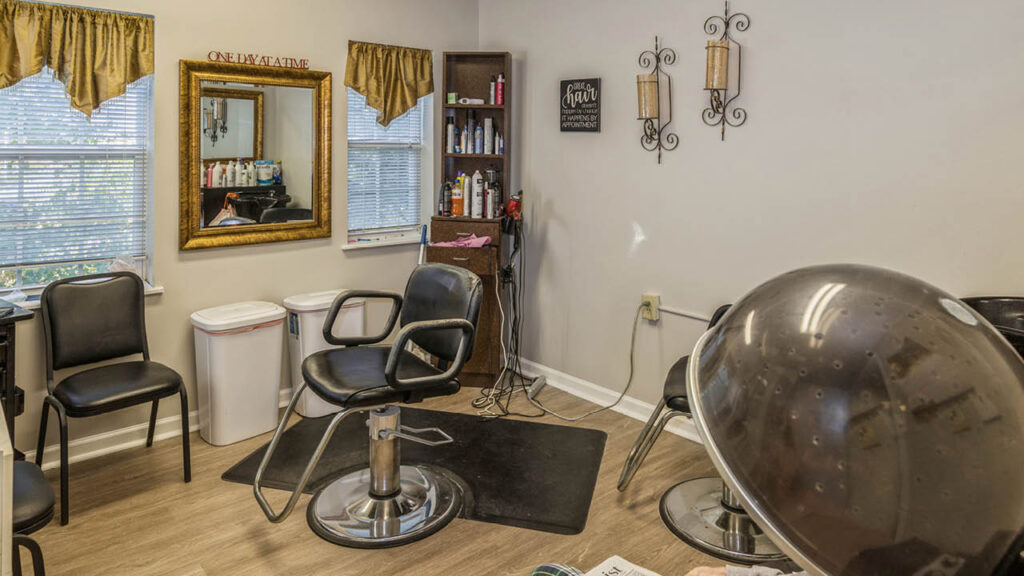 A small hair salon with black styling chairs, a wall mirror, hair products on a shelf, decorative wall sconces, a trash bin, and a hair dryer hood, illuminated by daylight from two windows with gold valances.