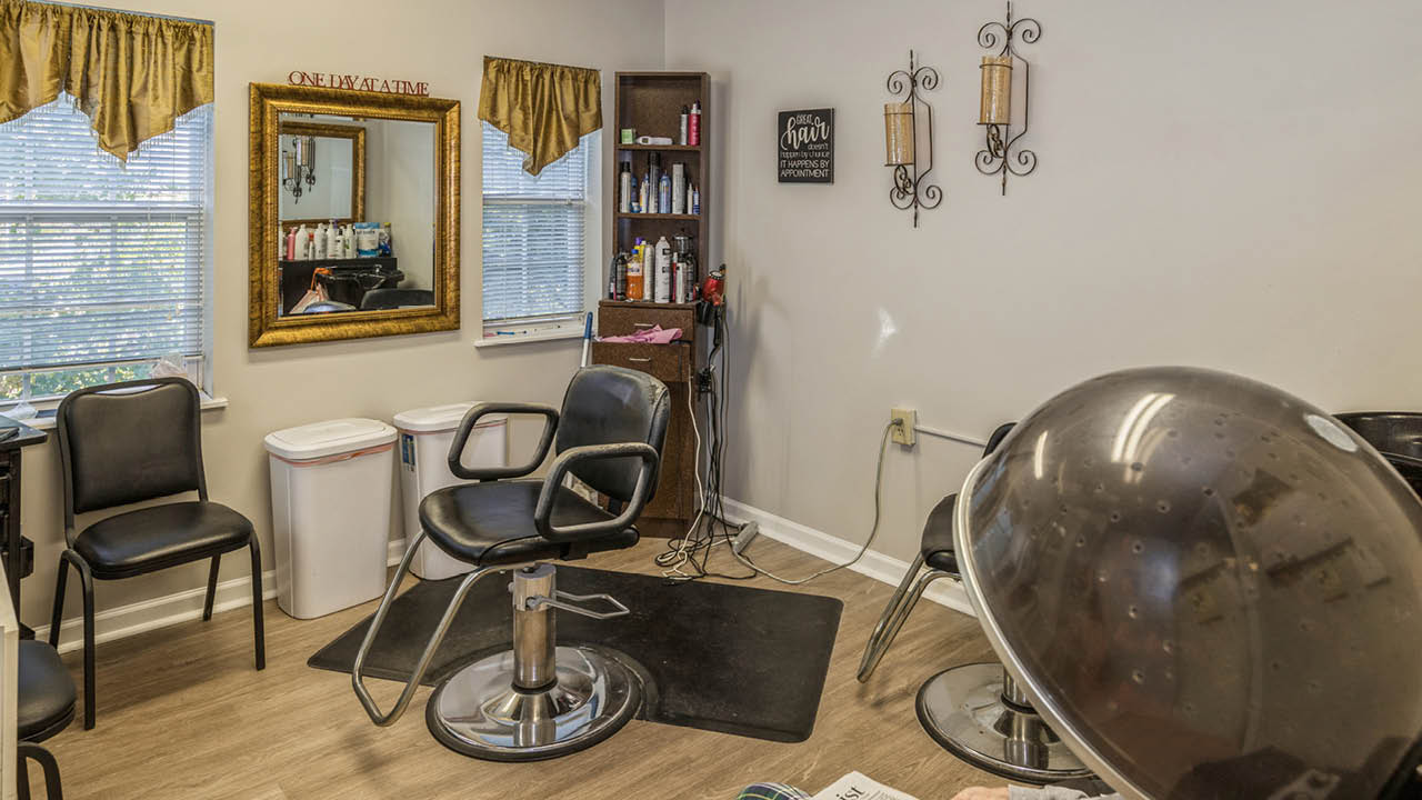 A small hair salon with black styling chairs, a wall mirror, hair products on a shelf, decorative wall sconces, a trash bin, and a hair dryer hood, illuminated by daylight from two windows with gold valances.