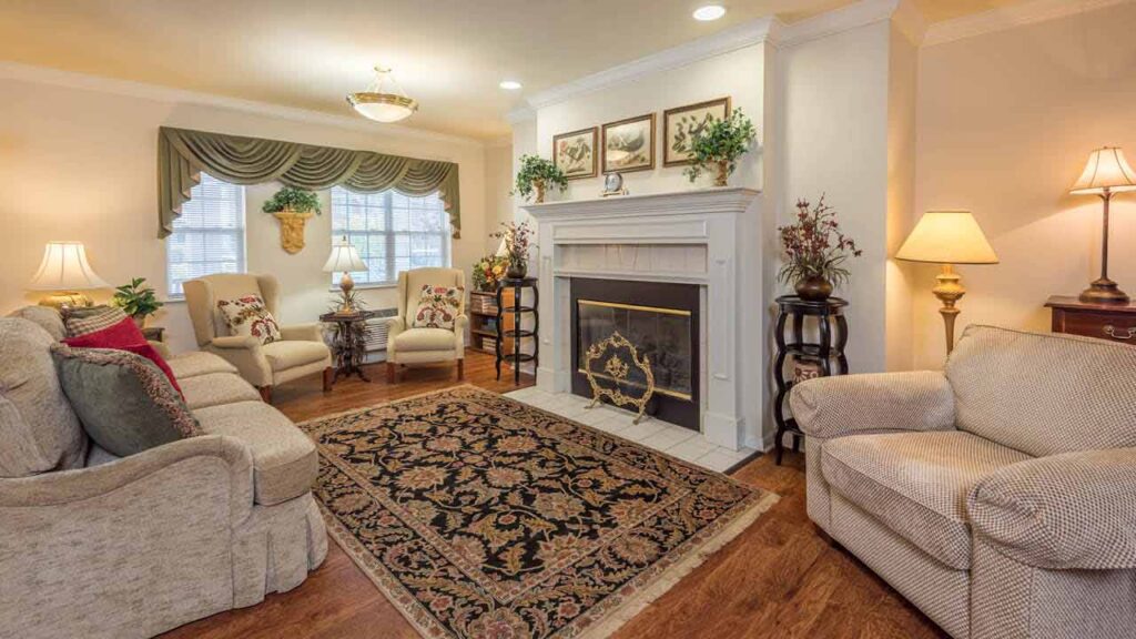 A cozy living room with beige sofas and armchairs, a fireplace with a decorative screen, a patterned rug, lamps, houseplants, and framed art above the mantel. Hardwood floors and a window with green curtains complete the space.