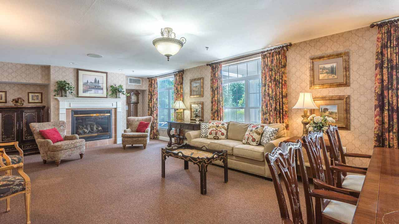 A cozy, traditionally decorated living room with beige walls, a fireplace, armchairs, a beige sofa, floral curtains, framed art, wooden dining table and chairs, lamps, and large windows letting in natural light.