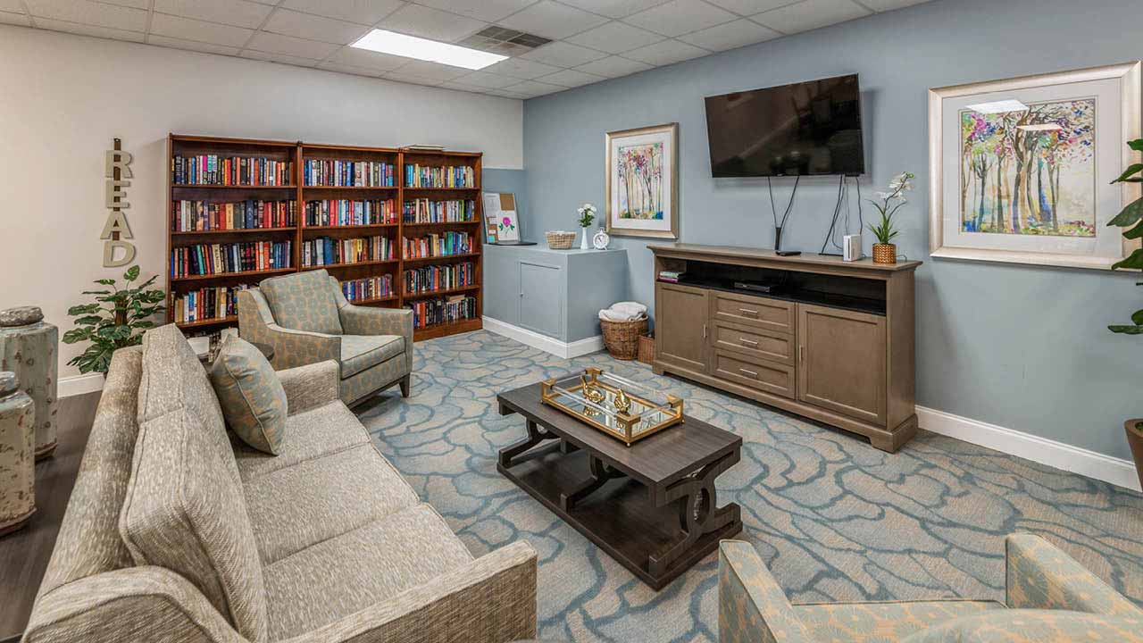A cozy reading room with a sofa, two armchairs, a coffee table with a chess set, a bookshelf filled with books, wall art, a TV mounted above a cabinet, and soft blue patterned carpet.