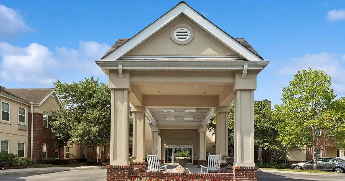 A covered entrance with columns leads to a brick-and-beige building. Two white rocking chairs sit at the front. Trees and blue sky with scattered clouds are visible in the background.