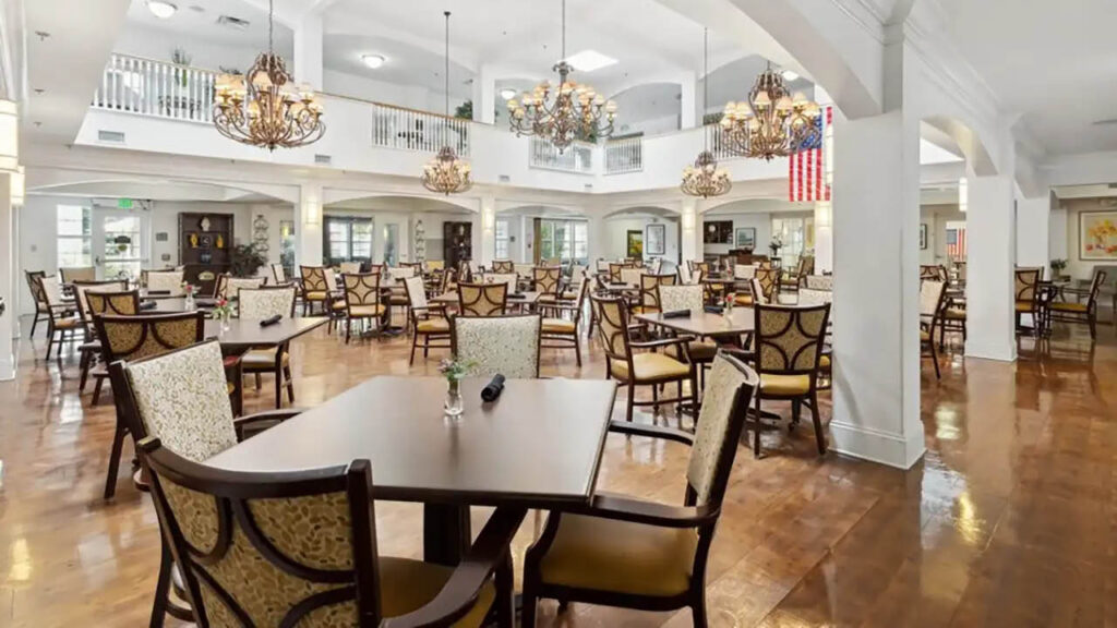 Spacious, elegant dining hall with many wooden tables and cushioned chairs, chandeliers hanging from the ceiling, polished floors, and an American flag on the wall. Large windows let in natural light.