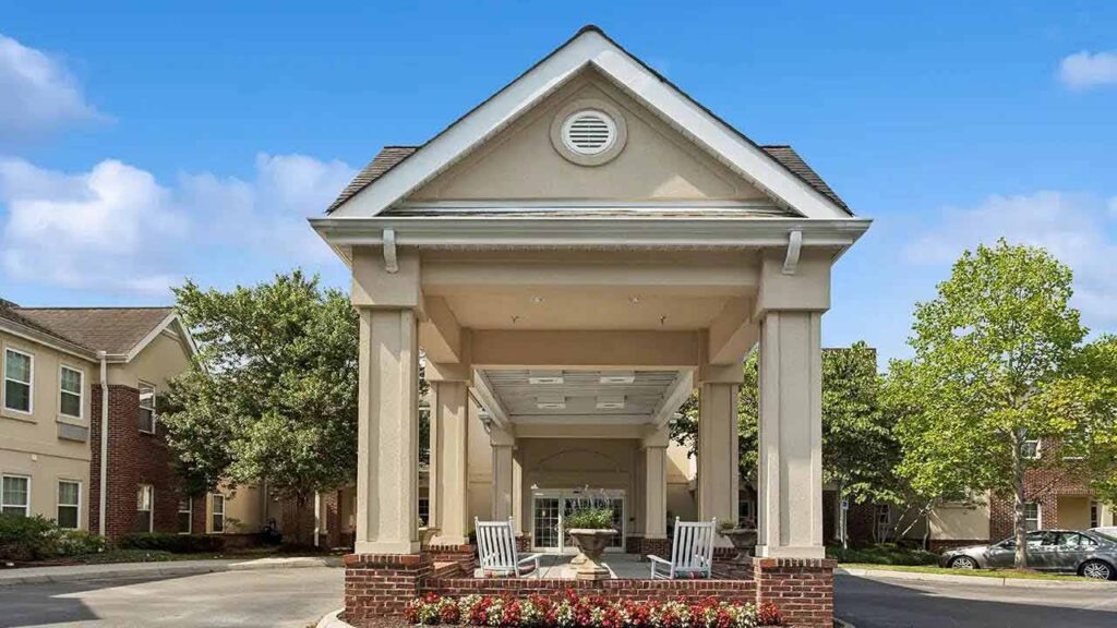 A covered entryway with columns leads to a building, flanked by rocking chairs and flower beds. The building has beige walls, brick accents, and is surrounded by trees under a blue sky.