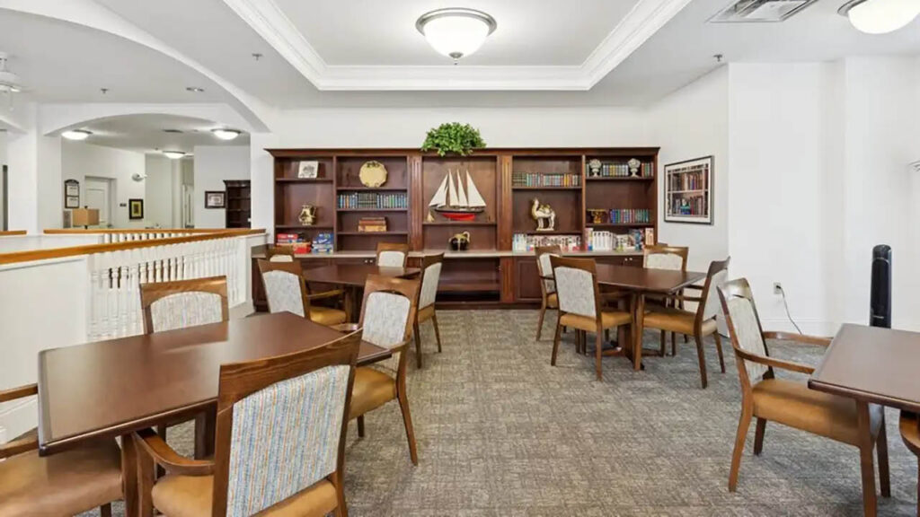 A well-lit room with wooden tables and chairs, carpeted floor, and a large bookshelf against the back wall displaying books, decorative objects, and a model sailboat. The space appears clean and organized.