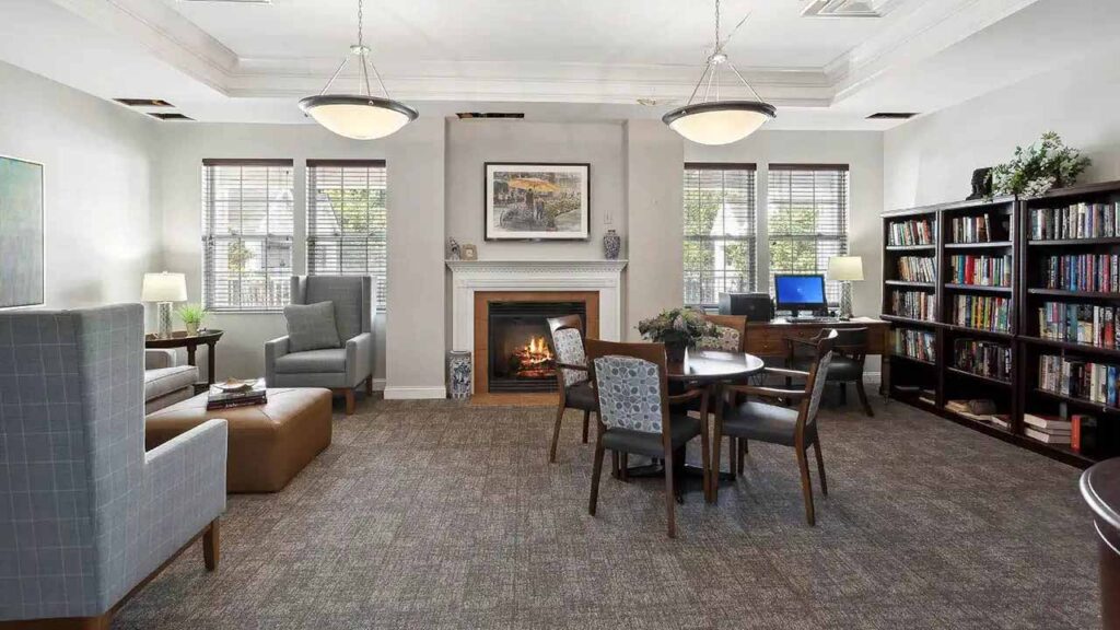A cozy library room with armchairs, a table with chairs, a fireplace, bookshelves filled with books, a desk with a computer, and large windows letting in natural light.