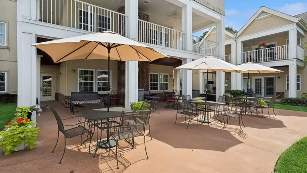 Outdoor patio area with metal tables and chairs under large beige umbrellas, surrounded by potted plants. A two-story building with white railing and balconies is in the background. The sky is clear and blue.