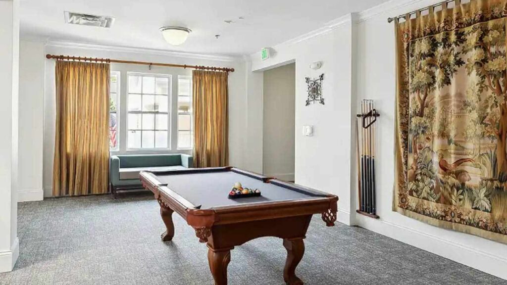 A well-lit room features a pool table with racked balls, a cue rack with pool cues, golden curtains, a window seat, and a decorative tapestry hanging on the wall.
