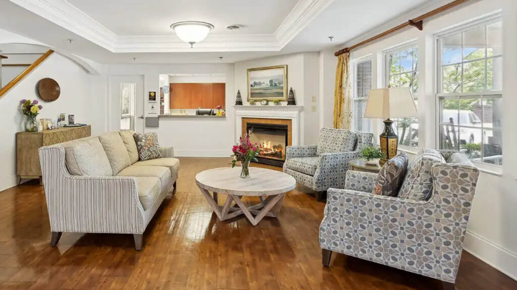 A cozy living room with a beige sofa, two patterned armchairs, a round wooden coffee table with flowers, a fireplace, hardwood floors, and large windows letting in natural light.