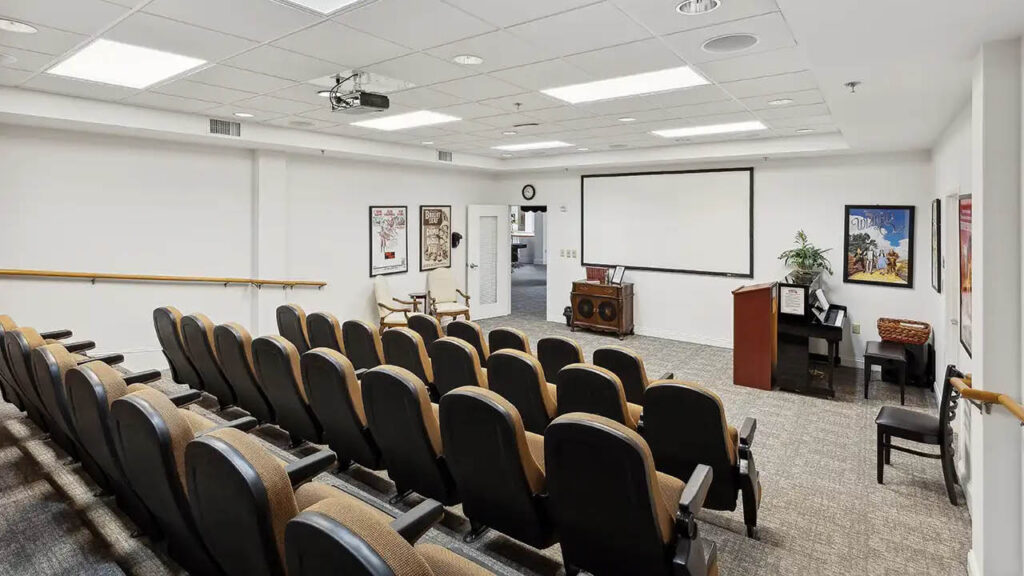 Small auditorium with rows of cushioned seats facing a projector screen, podium, piano, wall art, and a stereo system in a brightly lit room with white walls and carpeted floor.