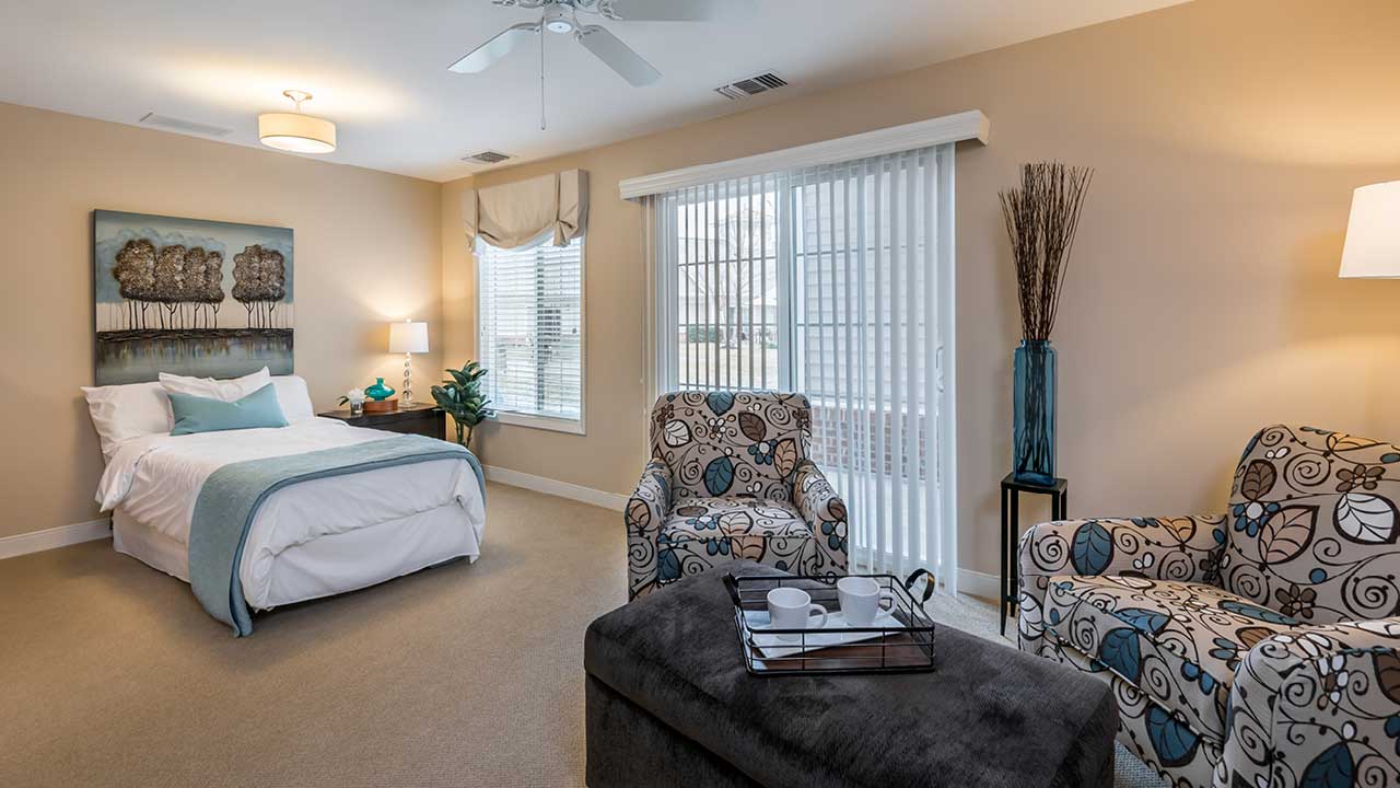 A cozy bedroom with a large bed, floral-patterned armchairs, a plush ottoman with a tray, and soft lighting. Large windows with blinds let in natural light, and beige walls create a warm atmosphere.