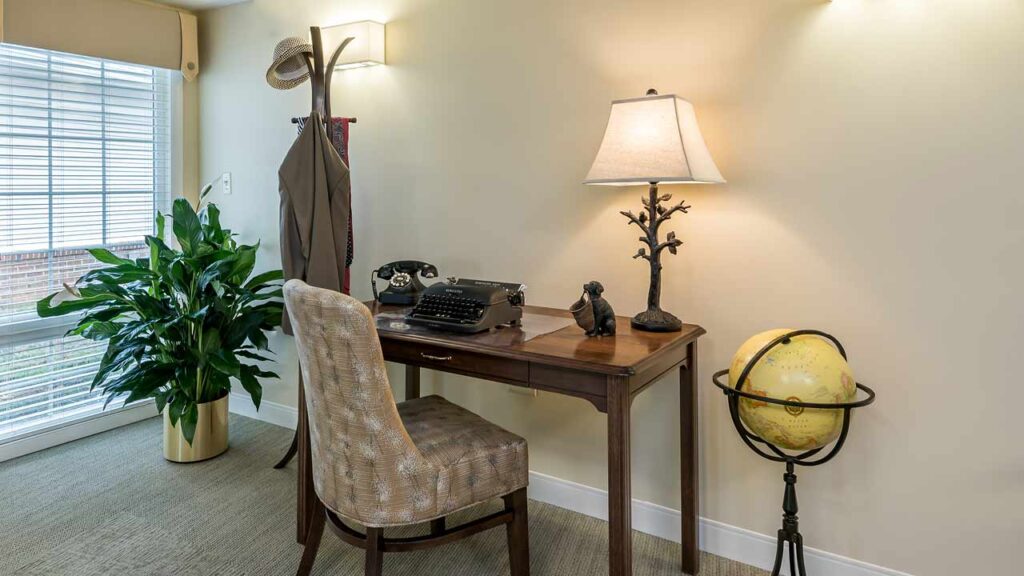 A small home office with a wooden desk, vintage typewriter, lamp, globe, and small sculptures. A patterned chair sits at the desk. A plant and coat rack are nearby, with natural light from a large window.