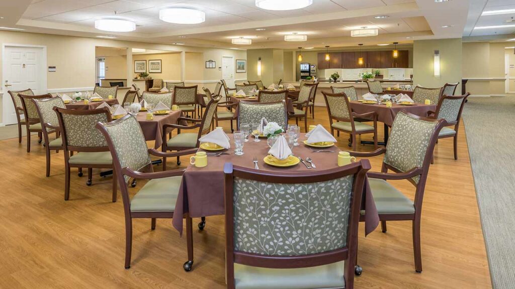 A spacious, well-lit dining room with round tables covered in brown tablecloths, neatly arranged chairs, and place settings including napkins, cups, and utensils, creating a welcoming atmosphere.
