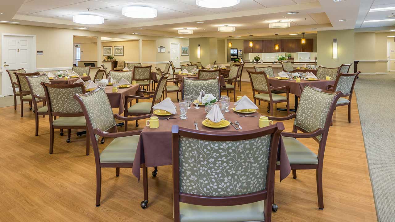 A spacious, well-lit dining room with round tables covered in brown tablecloths, neatly arranged chairs, and place settings including napkins, cups, and utensils, creating a welcoming atmosphere.