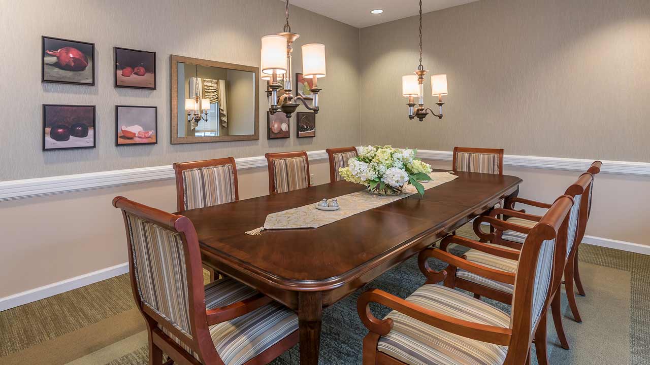 A dining room with a polished wooden table, eight striped upholstered chairs, floral centerpiece, wall art, a mirror, and two chandeliers hanging from the ceiling. The room has neutral-toned walls and carpet.