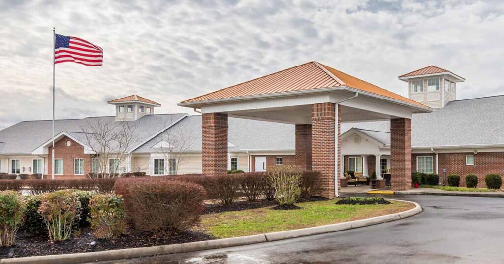 A brick building with a covered entrance, surrounded by neatly trimmed bushes. An American flag flies on a tall pole, and the sky is partly cloudy. The scene appears to be a residential or care facility.