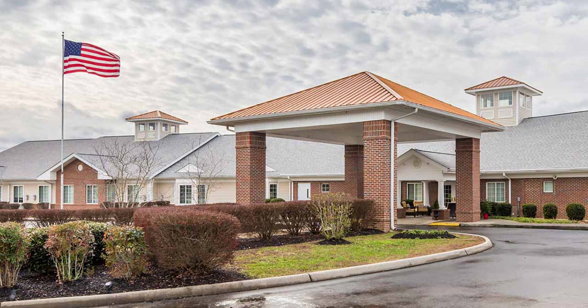 A brick building with a covered entrance, surrounded by neatly trimmed bushes. An American flag flies on a tall pole, and the sky is partly cloudy. The scene appears to be a residential or care facility.