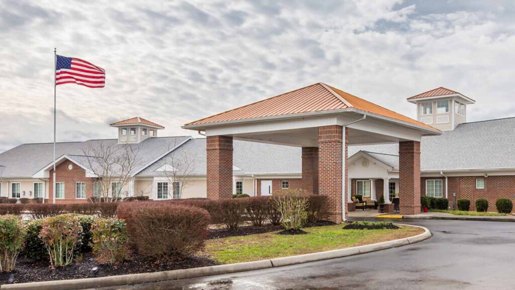 A large brick building with a covered entrance, surrounded by trimmed bushes and a circular driveway. An American flag flies on a tall flagpole, and the sky is partly cloudy above.